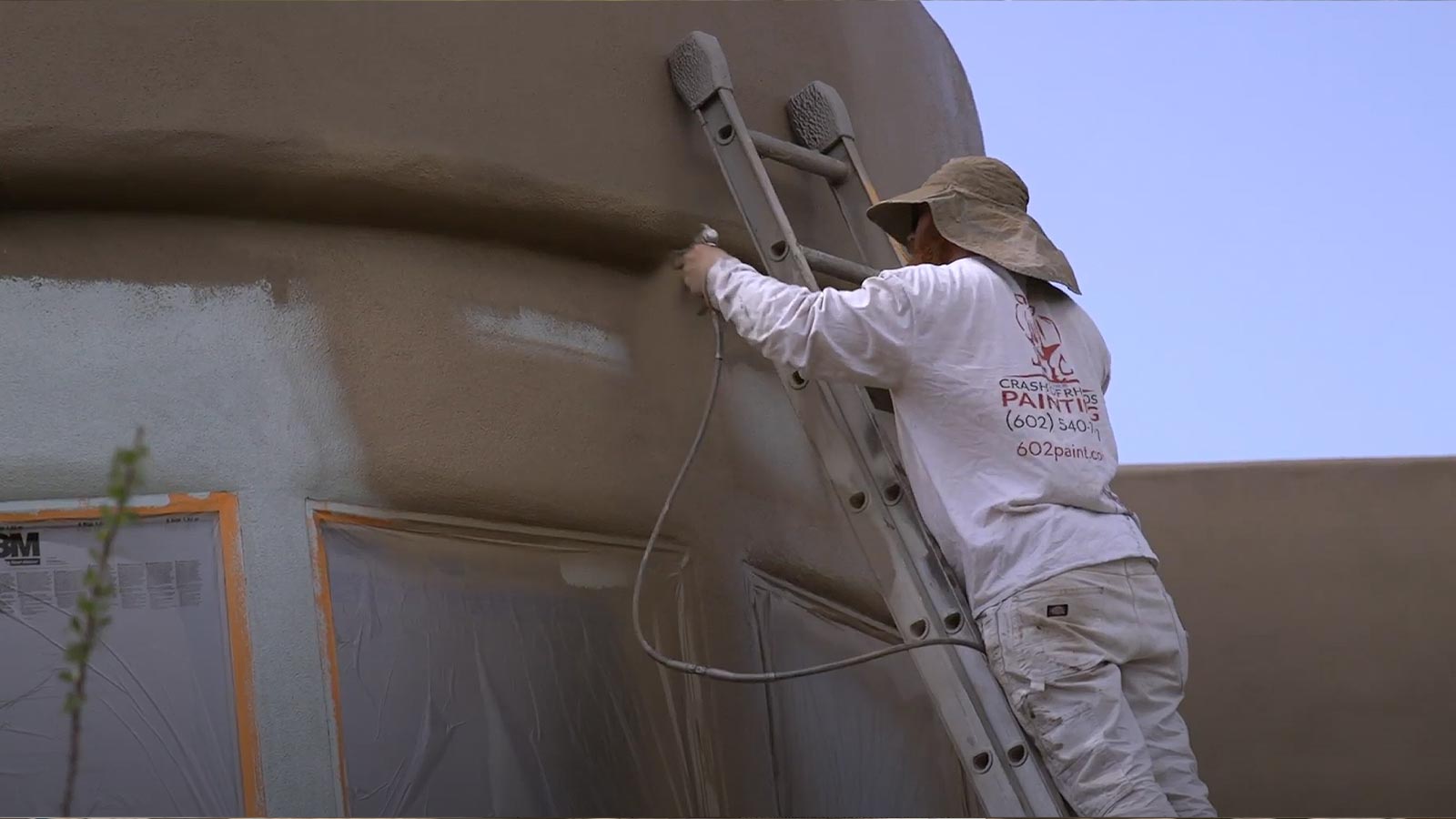 Crash of Rhinos painter spraying an exterior stucco wall from a ladder