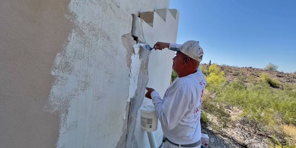 Person applying white paint to a textured wall outdoors.